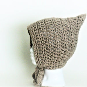 Może przedstawiać: Brązowy, szydełkowy czepek dla niemowlęcia z ostrym kapturem i długim sznurem. Czepek jest prezentowany na białej głowie manekina.