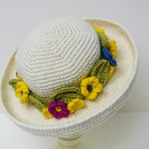 Puede incluir: Un sombrero de sol blanco de ganchillo con ala y una banda de flores de ganchillo amarillas, azules y verdes.