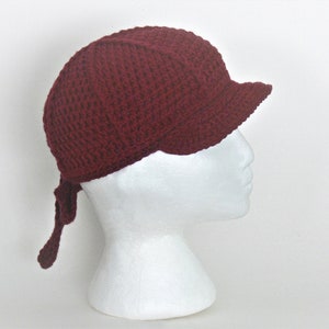 Può includere: Un cappello all'uncinetto color borgogna con visiera e un lungo laccio. Il cappello è su una testa di manichino bianca.