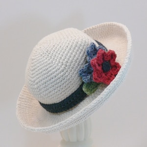 May include: A white crocheted sun hat with a wide brim and a navy blue ribbon. The hat is decorated with a cluster of crocheted flowers in shades of red, blue, and green.
