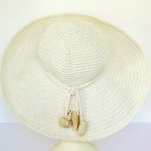 May include: A white crocheted sun hat with a wide brim and a knotted tie. The hat is decorated with small white seashells.