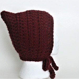 Op de afbeelding: Een bordeauxrode gebreide muts met capuchon en een lange strik. De muts is gemaakt met een textuur-steek en heeft een puntige capuchon.
