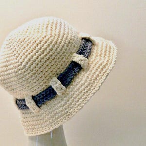 May include: Cream-colored crochet hat with a brim and a rounded crown. A dark blue band with white accents encircles the hat. The hat is made of a textured yarn, and the brim is slightly curved.