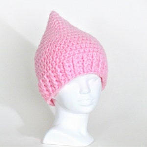 May include: A pink crocheted hat with a pointed top. The hat is on a white mannequin head.