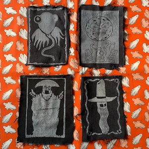 May include: Four black fabric patches with white designs. The designs include a creature with tentacles, a moon with a face, a person with a hat and a beard, and a person with a hat and a beard.