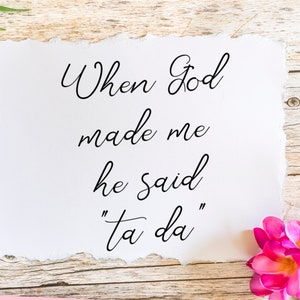 Può includere: Un foglio di carta bianco con il testo "When God made me he said "ta da" scritto in corsivo nero. La carta è su una superficie di legno con un fiore rosa nell'angolo in basso a destra.