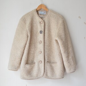 May include: A beige, fuzzy jacket with a round collar and decorative buttons. The jacket has two front pockets and a light brown trim. The buttons are a mix of clear and iridescent. The jacket is hanging on a wooden hanger.