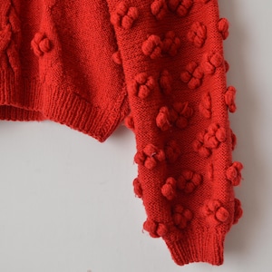 Könnte beinhalten: Ein leuchtend roter Strickpullover mit einem strukturierten Noppenmuster an Ärmel und Körper. Das Kleidungsstück hat einen gerippten Saum und gerippte Bündchen sowie eine dichte Anordnung von erhabenen, abgerundeten Strickclustern auf der gesamten Oberfläche.