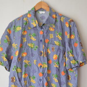 May include: A short-sleeved button-down shirt with a blue and white checkered pattern. The shirt is adorned with colorful illustrations of peaches, grapes, and other floral elements. The shirt is hanging on a wooden hanger.