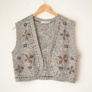 May include: A gray knitted vest with a floral pattern in shades of brown and blue. The sleeveless vest is hanging on a wooden hanger against a white background. The vest has a V-neck design.