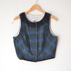 May include: A vintage plaid vest in shades of blue and green, with a black lace collar and button-up front. The vest is hanging on a wooden hanger against a white background. The garment has a fitted silhouette.