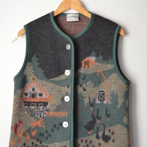 May include: A vintage, button-down vest with a rural landscape design. The sleeveless garment features a dark gray upper section and a green and tan lower section with images of houses, trees, and animals. The vest is hanging on a wooden hanger.