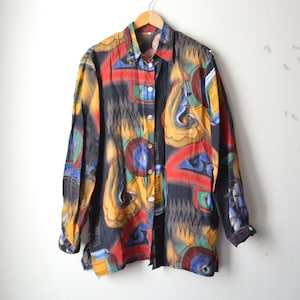 May include: A long-sleeved button-down shirt featuring a black base with a vibrant, multi-coloured abstract print. The design includes swirling shapes and geometric patterns in shades of yellow, red, blue, and green, with a standard collar and cuffs.