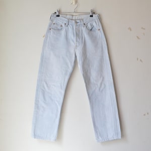 Pode incluir: Calças de ganga azul claro penduradas num cabide de metal. As calças de ganga têm um design clássico de cinco bolsos com fecho de botão e zíper. A ganga parece desbotada, dando às calças um visual vintage.