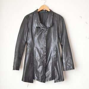 May include: A black, long-sleeved jacket with a unique collar design, hanging on a wooden hanger. The jacket has a button closure and a slightly flared bottom. The material appears to have a textured, possibly leather-like finish.