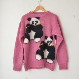 May include: A pink fluffy jumper with a black and white panda bear design. The panda bears are holding bamboo sticks.