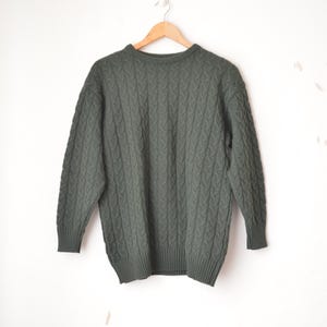 Peut inclure: Un pull vert foncé en tricot torsadé est suspendu à un cintre en bois contre un mur blanc. Le pull a un col rond et des manches longues. Le motif de tricot est un motif torsadé répétitif. Le vêtement semble être fait d'une matière épaisse et chaude.