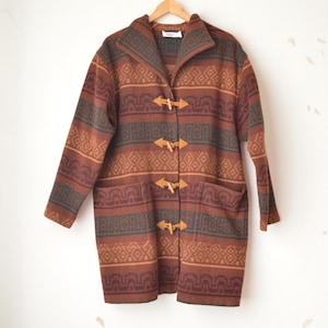 May include: A long, patterned coat in shades of brown, rust, and teal, with a geometric design. The coat features wooden toggle closures and two front pockets. It is hanging on a wooden hanger.