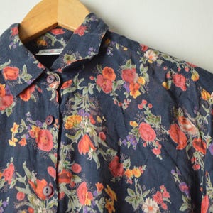 May include: A dark navy blue button-down shirt featuring a vibrant floral pattern with red, orange, yellow, and purple flowers and green leaves. The shirt has a classic collar, long sleeves, and matching dark navy blue buttons down the front.