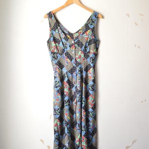 Peut inclure: Robe vintage sans manches avec un col en V et une fermeture boutonnée. La robe présente un motif patchwork avec des motifs floraux et géométriques dans des tons de bleu, noir et rouge. Suspendue à un cintre en bois contre un mur blanc.