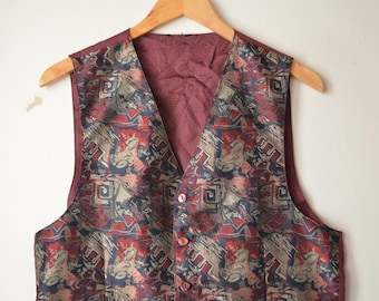 May include: A vintage vest with a burgundy interior and a patterned exterior. The vest's exterior features a mix of red, blue, and tan geometric shapes and floral designs. It is buttoned up and hanging on a wooden hanger.