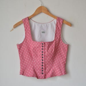 May include: A pink sleeveless top with a square neckline and a white lining. The top is adorned with small white polka dots and features a row of dark buttons down the front. It hangs on a wooden hanger.