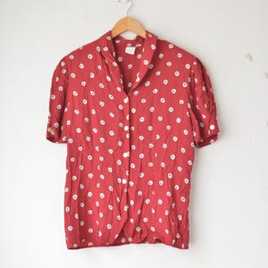 May include: Red short-sleeved button-down shirt with white polka dots. The shirt has a collared neckline and is hanging on a wooden hanger against a white wall. The shirt is made of a lightweight fabric.