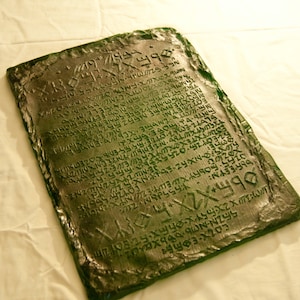 May include: A green stone tablet with an inscription of an ancient language. The tablet is rectangular and has a rough, textured surface.
