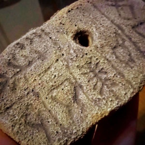 May include: A close-up of a gray, textured, rectangular object with a hole in the center. The object has a rough, uneven surface and appears to be made of concrete or stone.