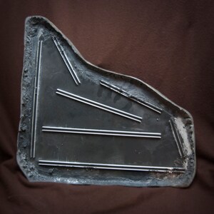May include: A gray metal car part with a textured surface and multiple parallel grooves. The part is shaped like a triangle with rounded edges.