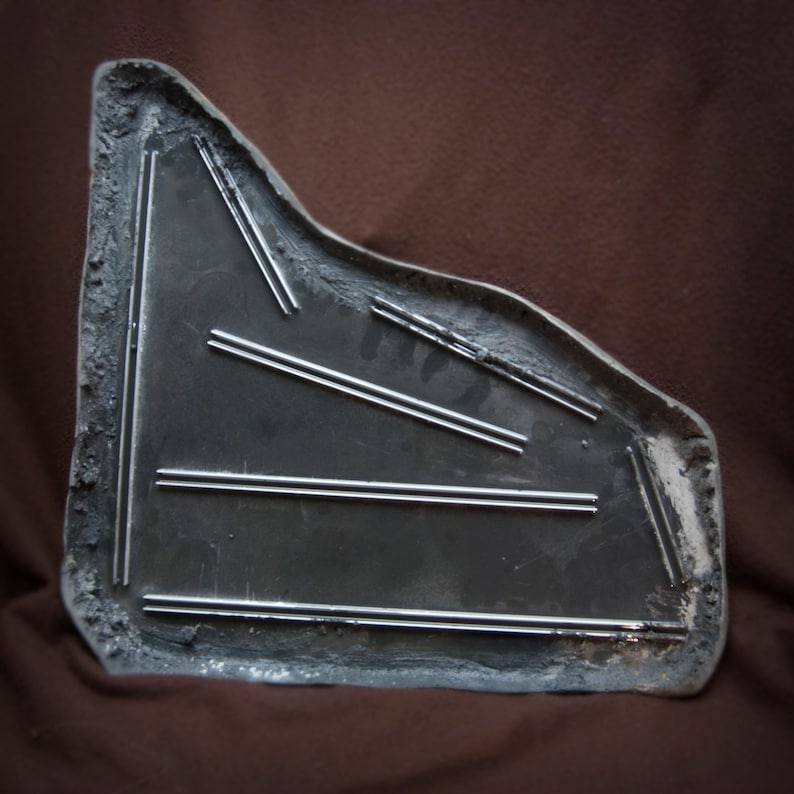 May include: A gray metal car part with a textured surface and multiple parallel grooves. The part is shaped like a triangle with rounded edges.