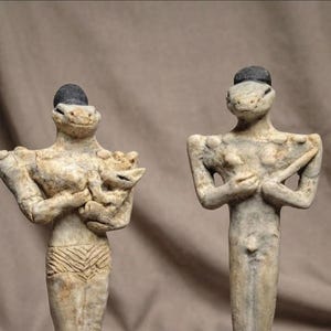 Può includere: Due statuette in argilla beige con teste di rettile e piccoli cappelli neri. La figura a sinistra tiene in braccio un piccolo animale, mentre quella a destra è in piedi con le braccia incrociate. Entrambe hanno superfici ruvide e torsi umani.