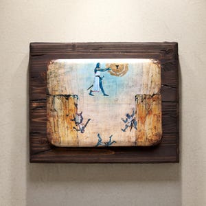 May include: A framed artwork with a medieval illustration. The artwork shows figures in blue and tan clothing, with a central figure holding a sword. The background is light blue and tan, framed in a dark brown wooden frame.