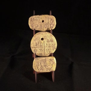 May include: Three round, yellow clay tokens with markings on them are displayed on a wooden stand. The tokens have holes in the center and are arranged vertically.