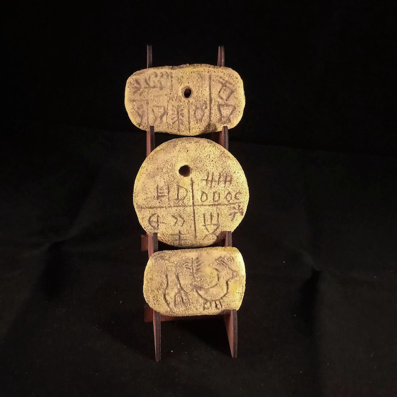 May include: Three round, yellow clay tokens with markings on them are displayed on a wooden stand. The tokens have holes in the center and are arranged vertically.