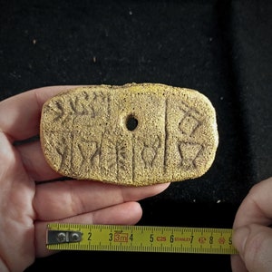 May include: A gold-colored clay tablet with cuneiform script, a type of ancient writing system. The tablet is rectangular and has a hole in the center.
