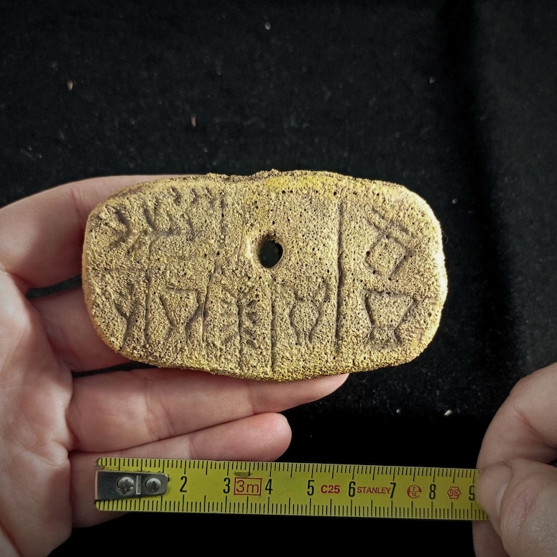 May include: A gold-colored clay tablet with cuneiform script, a type of ancient writing system. The tablet is rectangular and has a hole in the center.