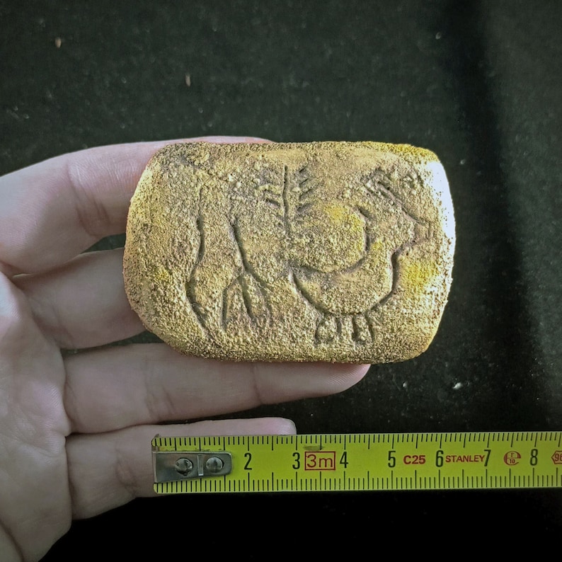 May include: A gold-colored rectangular stone with a carved design of a bird and a plant. The stone is about 3 inches long.