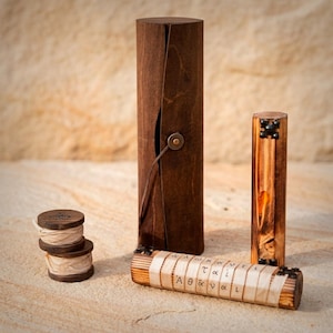 Op de afbeelding: Een verzameling houten items, waaronder een hoge, donkerbruine houten doos met een leren riemsluiting, een kleinere houten cilinder met decoratieve metalen studs en een houten cilinder met Griekse letters. Er zijn ook spoelen met lint.