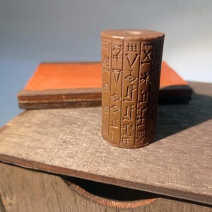 Può includere: Un oggetto cilindrico marrone con testo antico scolpito. È posto su una superficie di legno, con un oggetto rettangolare sullo sfondo. Probabilmente un manufatto storico o un oggetto decorativo.