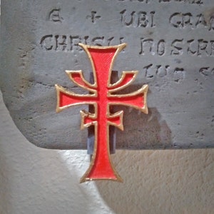 May include: A red and gold cross with a decorative design. The cross is mounted on a gray stone surface with the text "E + UBI GRA" and "CHRISTI NOS TR" visible.