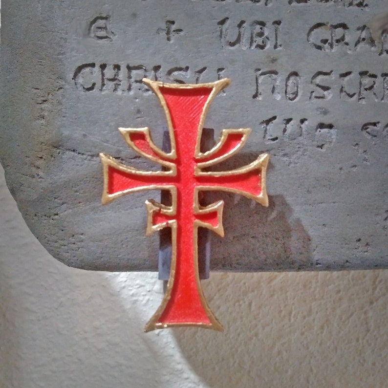 May include: A red and gold cross with a decorative design. The cross is mounted on a gray stone surface with the text "E + UBI GRA" and "CHRISTI NOS TR" visible.