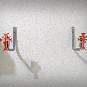 May include: Two silver wall hooks with a red and gold cross design. The hooks are mounted on a white wall.