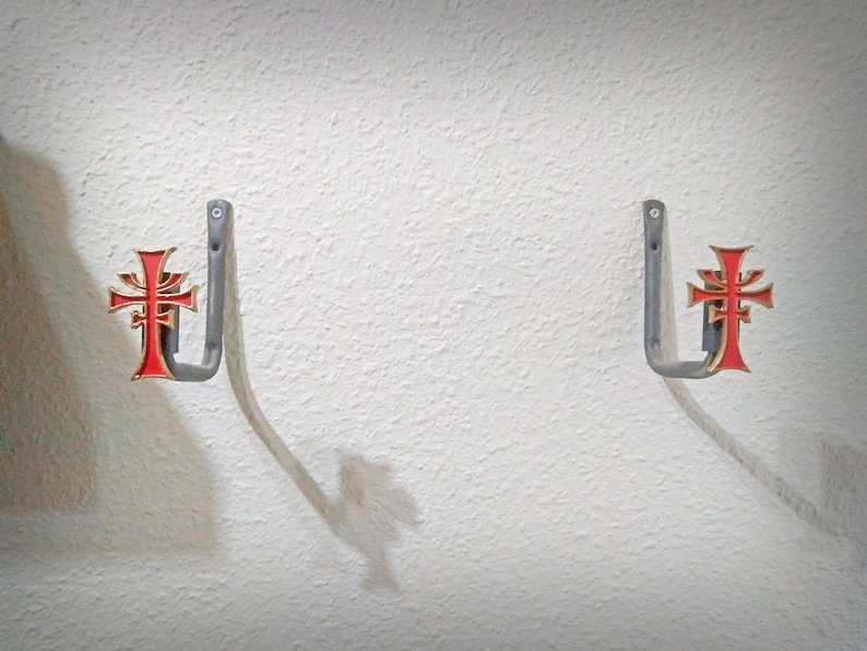 May include: Two silver wall hooks with a red and gold cross design. The hooks are mounted on a white wall.
