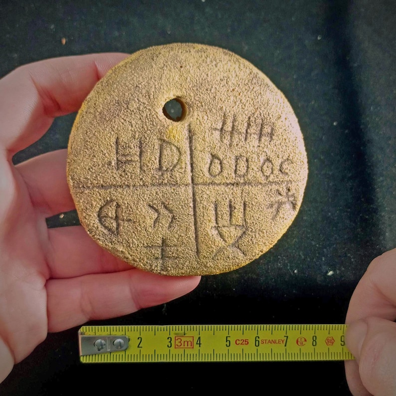 May include: A round, gold-colored, ceramic token with a hole in the center. The token is divided into four sections, each with a different inscription. The inscriptions are: "HD", "000C", "4", and "11". The token measures approximately 2 inches in diameter.
