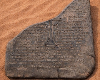 May include: A dark gray, irregularly shaped stone slab featuring an engraved cross in the center and extensive Latin text inscribed across its surface. The stone has a weathered, textured appearance with visible cracks and natural variations in its surface.