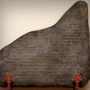 May include: A dark gray stone with gold lettering and two red crosses. The stone has a rough, uneven surface and is inscribed with a Latin text. The text reads: "ST ARA SE SURGE DOONE KERONOM BARFUM QUI HABITANZ LAUDATU ZG IM SECULA zu Deus as bellu SOLO HAG QT SA SACRAD Conted in Lace PARGUSEAS CRESCERLIS LETAG CRUCIAZA LEBAR CHRISELO DEE TOUS UBIQUS CIS POR ISSU in SIC AQUAN RUCIALA MABGBIC FOTZER TERE METERRAE FUGILE REX DEORUM MOSEROKE In AEDE LUA DOOMORR SASCUILORGIO SU RAFTIGED: 0017 DUCATO DE AD Quem moue: PL SOOS LACAS AD EGIPELIR SOLES CORIBUS VERS SALS Sea BUST GRADALIS GUAE constat SanCURS 1850 CHRISTI NOGERI PORRATMEL IN ACUSATUM LAD SOLIS OUTRAS ORIS SADIS SANCE"