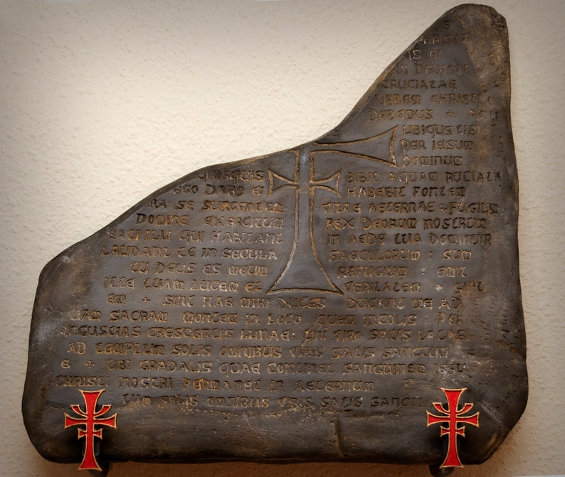 May include: A dark gray stone with gold lettering and two red crosses. The stone has a rough, uneven surface and is inscribed with a Latin text. The text reads: "ST ARA SE SURGE DOONE KERONOM BARFUM QUI HABITANZ LAUDATU ZG IM SECULA zu Deus as bellu SOLO HAG QT SA SACRAD Conted in Lace PARGUSEAS CRESCERLIS LETAG CRUCIAZA LEBAR CHRISELO DEE TOUS UBIQUS CIS POR ISSU in SIC AQUAN RUCIALA MABGBIC FOTZER TERE METERRAE FUGILE REX DEORUM MOSEROKE In AEDE LUA DOOMORR SASCUILORGIO SU RAFTIGED: 0017 DUCATO DE AD Quem moue: PL SOOS LACAS AD EGIPELIR SOLES CORIBUS VERS SALS Sea BUST GRADALIS GUAE constat SanCURS 1850 CHRISTI NOGERI PORRATMEL IN ACUSATUM LAD SOLIS OUTRAS ORIS SADIS SANCE"