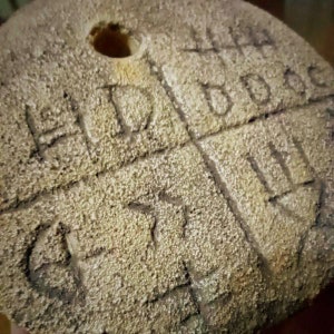 May include: A close-up of a wooden disc with a hole in the center. The disc is covered in a rough, textured surface and has various markings carved into it.