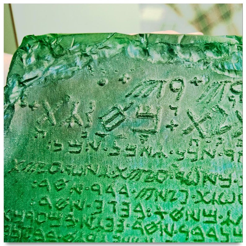 May include: A close-up of a green stone tablet with ancient writing carved into its surface. The writing is in a language that is difficult to decipher, but it appears to be a form of cuneiform script. The tablet is likely from an ancient civilization, and it is a fascinating artifact of history.